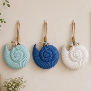 May include: Three nautilus shell wall decorations in light blue, dark blue, and white. Each shell features a textured spiral design, a jute rope loop for hanging, and a small decorative starfish accent attached to the top of the rope.