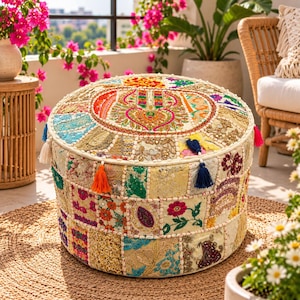 Puede incluir: Un puf redondo y bajo con un diseño de patchwork de paneles de tela bordados en colores vibrantes que incluyen rosa, naranja, azul y dorado. La superficie está decorada con intrincados patrones florales, lentejuelas y pequeñas borlas.