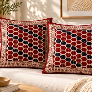 May include: Two square throw pillows featuring a repeating geometric hexagon pattern in deep red and dark blue. The central design is framed by a wide, cream-colored border with intricate, small-scale dark brown patterns and a solid red edge.
