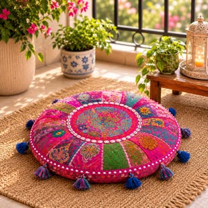 May include: A round floor cushion featuring a vibrant patchwork of pink, magenta, green, and orange fabrics with intricate embroidery. The cushion is accented with a border of small white shells and finished with blue tassels along the outer edge.