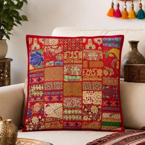 May include: A square throw pillow featuring a patchwork design of red fabric panels. Each panel is decorated with intricate gold-tone embroidery, sequins, and colourful floral or geometric patterns. The pillow has a consistent red border.