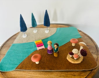 Puede incluir: Un tapete de juego de fieltro con un río, un puente y árboles. Figuras de madera de un hombre y una mujer están cerca del puente. También hay setas de madera y setas de fieltro sobre una base de madera. La escena está sobre una mesa redonda de madera.