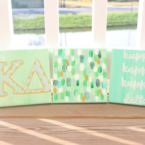 May include: Three square canvases featuring a light green background. The first canvas displays the Greek letters Kappa Delta in gold with a white outline. The second canvas has a pattern of small green, white, and orange brushstrokes. The third canvas features the words kappa kappa kappa delta in white script.