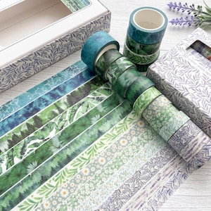 May include: A set of 30 rolls of washi tape featuring various green and purple botanical, floral, and forest patterns. The collection includes rolls with depictions of leaves, trees, daisies, and lavender, presented in a patterned box.