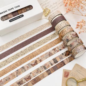 May include: A set of thin washi tape rolls in a white box labeled WASHI TAPE THIN handwritten text. The tapes feature various vintage-inspired designs including brown cursive script, sepia-toned newspaper clippings, and detailed butterfly illustrations.