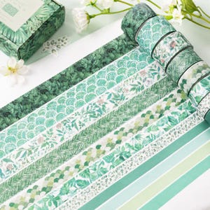 May include: A set of decorative washi tape rolls featuring various green botanical and geometric patterns, including fern leaves, scalloped scales, floral motifs, herringbone textures, and simple stripes, arranged in a row on a white surface.