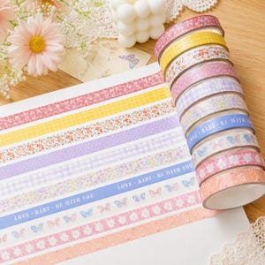 Op de afbeelding: Een stapel van tien rollen decoratieve washi-tape met diverse patronen, waaronder bloemenprints, geel ruitpatroon, paars gingham en een blauwe tape met de tekst LOVE BABY BE WITH YOU en vlinderillustraties op een wit vel papier.