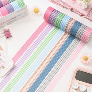 May include: A set of twelve rolls of washi tape featuring various grid and gingham patterns in pink, purple, green, blue, and brown. The rolls are displayed in a white box labeled Basic Washi Tape and unrolled in parallel lines on a surface.