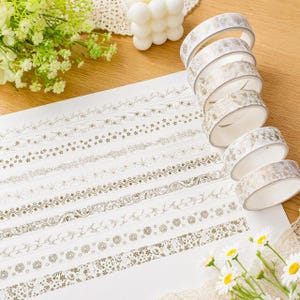 May include: A stack of six rolls of decorative adhesive tape featuring gold foil floral and geometric patterns. The tape is displayed alongside a white sheet showing the patterns unrolled, with each strip showcasing unique botanical designs.