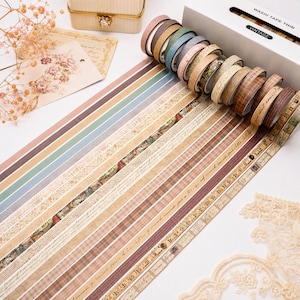 May include: A set of thin washi tape rolls in a white box labeled WASHI TAPE THIN VINTAGE. The rolls feature various patterns including solid colors, plaid, script, and floral designs in shades of brown, beige, blue, and green, displayed in a row.