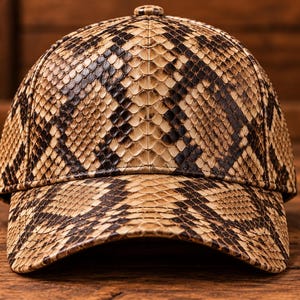 Può includere: Un cappellino da baseball con un motivo dettagliato in pelle di serpente nei toni del beige, marrone chiaro e marrone scuro. Il cappellino presenta una corona strutturata e una visiera curva, con la trama a scaglie su tutta la superficie.