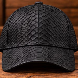 Könnte beinhalten: Eine schwarze Baseballkappe mit einer strukturierten Schlangenledermusterung auf der gesamten Oberfläche. Die Kappe hat eine strukturierte Krone, einen gebogenen Schirm und einen kleinen Knopf in der Mitte oben. Das Material zeigt ein Muster.