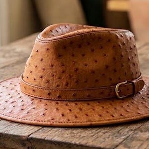 May include: A tan fedora hat with a textured ostrich-skin pattern featuring small, dark, recessed spots across the crown and brim. The hat includes a matching tan leather band around the base of the crown, secured with a small, metallic buckle.
