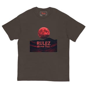 Puede incluir: Una camiseta de manga corta de color marrón oscuro con un estampado gráfico de una luna roja sobre un paisaje oscuro y texturizado. El texto "RULEZ" aparece en letras rojas grandes sobre el texto rojo más pequeño "Strange Dayz".