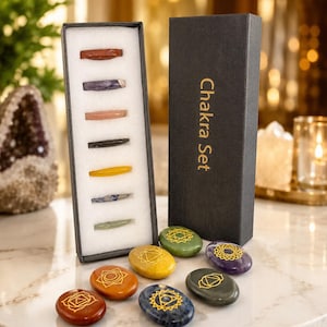 May include: A black rectangular box labeled Chakra Set containing seven slender polished stones in a white foam insert, accompanied by seven oval-shaped stones of various colors, each engraved with a unique gold-colored geometric symbol.
