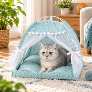 May include: A light blue pet tent with a white polka dot pattern, featuring white sheer curtains tied at the sides with ribbons and a white pom-pom trim along the top edge. A grey tabby cat is resting inside on a matching cushioned base.