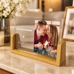 Può includere: Una cornice portafoto rettangolare in acrilico trasparente con base in legno chiaro. Il pannello in acrilico presenta il testo stampato LOVE YOU FOREVER, un piccolo cuore e i nomi Rose & Joel, mostrando la foto di una coppia con una scintilla.