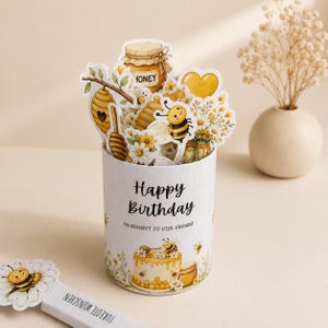 Könnte beinhalten: Ein zylindrischer Behälter mit dem Text Happy Birthday und Du bringst so viel Freude, verziert mit Illustrationen von Kuchen, Bienen und Blumen. Darin befinden sich verschiedene ausgestanzte Papieraufsteller mit Honiggläsern und Bienen.