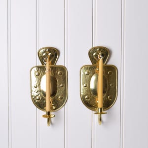 May include: A pair of polished brass wall sconces featuring a hammered texture on the backplate, a central oval protrusion, and a small circular candle cup at the base. Each sconce holds a single slender, light beige taper candle.