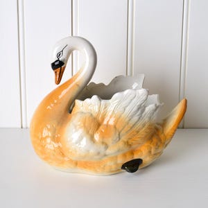 May include: A ceramic swan-shaped planter or decorative bowl featuring a glossy finish. The swan has a white neck and head with a black and orange beak, transitioning into a textured body with orange and white plumage and a hollow centre.