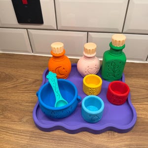 Pode incluir: Um conjunto de objetos de brincar em silicone colorido sobre um tabuleiro roxo, incluindo uma garrafa laranja com uma lua, uma garrafa rosa com um coração, uma garrafa verde com uma folha, uma taça azul com colher e três copos pequenos.