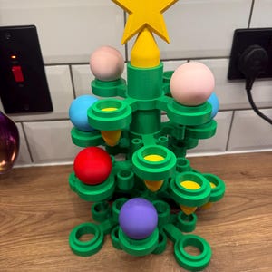 May include: A green plastic Christmas tree structure featuring a yellow star topper. The tree has multiple circular holders containing various coloured spheres, including pink, blue, red, and purple, along with small yellow cone-shaped ornaments.