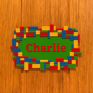 Può includere: Una targa rettangolare verde con il nome Charlie scritto in lettere rosse. La targa è circondata da una cornice costruita con piccoli mattoncini di plastica a incastro nei colori primari rosso, blu, giallo e verde.