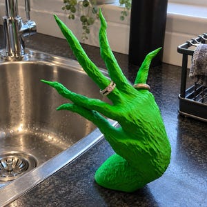 May include: A bright green, textured hand-shaped jewellery display stand with long, pointed fingers. The stand features a ring with a small pink stone on the middle finger and a plain metallic band on the ring finger, showcasing a detailed surface.