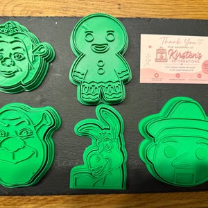 May include: Five green plastic cookie cutters featuring character faces and shapes, including a princess, a gingerbread person, an ogre, a donkey, and a puppet. A small rectangular card with the text Kirsten's 3D Creations is included.