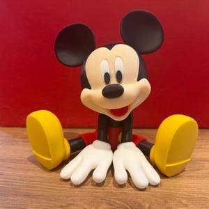 May include: A Mickey Mouse figurine in a seated position, featuring a black head with large round ears, a cream-coloured face, white gloves, red shorts, and bright yellow shoes. The character has a wide, open-mouthed smile and black limbs.