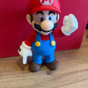 May include: A plastic figurine of the character Mario, featuring a red cap with a white M emblem, a red shirt, blue overalls with yellow buttons, white gloves, and brown shoes. The figure is posed with one hand raised and one hand pointing downward.