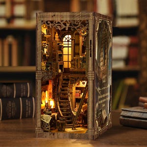 May include: A wooden book nook insert featuring a miniature library scene with a curved staircase, shelves filled with tiny books, and warm interior lighting. The exterior side panel displays a printed graphic with text about knowledge and books.