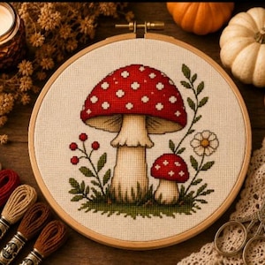 Pode incluir: Um bastidor de bordado circular de madeira contendo um padrão de ponto cruz de dois cogumelos vermelhos com pintas brancas, uma pequena flor branca e folhagem verde em tecido creme. O design está centralizado na moldura.