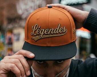 May include: A burnt orange baseball cap with a flat black brim. The front features the word "Légendes" embroidered in a gold script font with a dark outline. A small gold embroidered graphic of two crossed baseball bats is on the side panel.