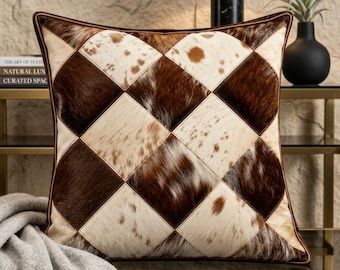 May include: A square throw pillow featuring a patchwork design of brown and white cowhide leather arranged in a diamond pattern. The pillow has a dark brown leather border and a textured, natural hair-on-hide finish with varied color tones.