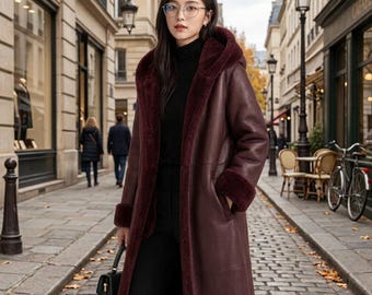 May include: A long, deep burgundy coat featuring a hood and a plush, matching faux fur lining visible along the front opening and cuffs. The coat is worn over a black turtleneck sweater and black trousers, paired with a small black handbag.
