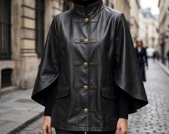 May include: A black leather cape jacket featuring a high collar, four gold-toned decorative buttons with toggle-style closures, and two front flap pockets. The garment has wide, open sleeves and a smooth finish, worn over a black long-sleeved top.