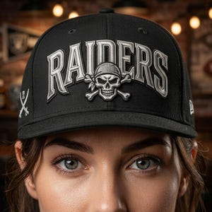 Könnte beinhalten: Eine schwarze Baseballkappe mit dem in silbernem Garn gestickten Wort RAIDERS auf der Vorderseite. Ein silberner Totenkopf mit Bandana und gekreuzten Knochen ist unter dem Text zentriert. Ein kleines weißes Emblem mit gekreuzten Schwertern ist seitlich.