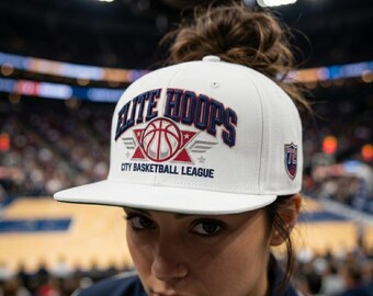 May include: A white snapback cap featuring the embroidered text ELITE HOOPS and CITY BASKETBALL LEAGUE in navy blue. The design includes a central basketball graphic with red star accents and wings, plus a small shield patch with the number 72 on the side.