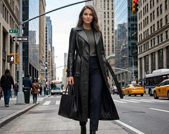 May include: A long black leather trench coat with a belted waist, worn over a dark grey sweater and dark trousers. The coat features a wide collar, long sleeves, and a structured fit. A black leather tote bag is carried in one hand.