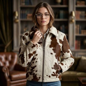 May include: A zip-up jacket featuring a brown and white cow print pattern, a structured collar, long sleeves with cuffs, and two side pockets. The garment is worn over a light-colored collared shirt and paired with blue denim jeans.