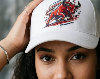 May include: A white baseball cap featuring an embroidered graphic of a charging red bull with dark blue and white accents. The design includes stylized motion lines and geometric shapes surrounding the bull, positioned on the front panel.