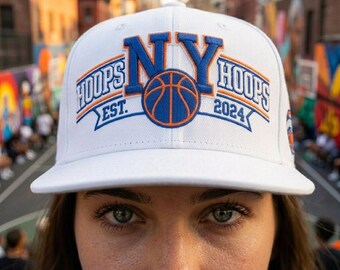 May include: A white baseball cap featuring embroidered text that reads NY HOOPS EST. 2024 with a central basketball graphic. The embroidery uses blue and orange thread. The cap has a structured crown and a flat brim, with a small logo on the side panel.