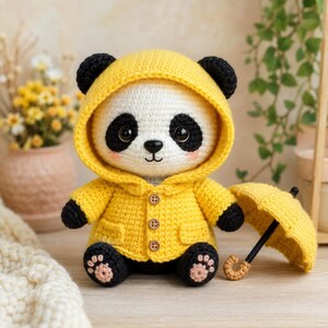 May include: A crocheted panda doll wearing a bright yellow hooded raincoat with three brown buttons and two pockets. The panda has black ears, black eye patches, and pink cheeks. It is holding a small yellow umbrella with a black handle.
