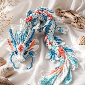 Puede incluir: Una figura de dragón tejida a ganchillo con un cuerpo largo y serpentino con escamas intrincadas en tonos blanco, azul claro y naranja coral. La cabeza tiene cuernos blancos, bigotes azules y ojos oscuros, con aletas a lo largo del cuerpo.