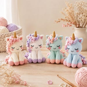 May include: Four crocheted unicorn plush toys arranged in a row. Each unicorn features a gold horn, a curly mane in pastel shades, closed eyes with eyelashes, and pink cheeks. The bodies are white, mint green, and light blue with pink hooves.