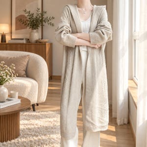 May include: A long, beige, open-front hooded cardigan featuring long sleeves with ribbed cuffs and a subtle vertical cable knit pattern along the front panels. The garment is worn over a white ribbed tank top and matching white trousers.