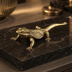 May include: A metallic gold-toned lizard figurine with a textured, scaly surface and a long tail, positioned in a crawling stance. The lizard features detailed eyes, a wide head, and four legs with visible claws, crafted with a realistic appearance.