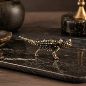 Può includere: Una piccola e dettagliata statuetta in metallo color bronzo che raffigura un ramarro cornuto. La scultura presenta un dorso ruvido e spinoso, una lunga coda e distintive protuberanze simili a corna sulla testa, in posizione di profilo.
