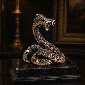 Op de afbeelding: Een metalen cobra-figuur in bronskleur in een aanvallende houding met een gespreide kap en een open bek die giftanden toont. De slang heeft gedetailleerde schubben en is bevestigd op een rechthoekige zwarte marmeren voet met witte aders.